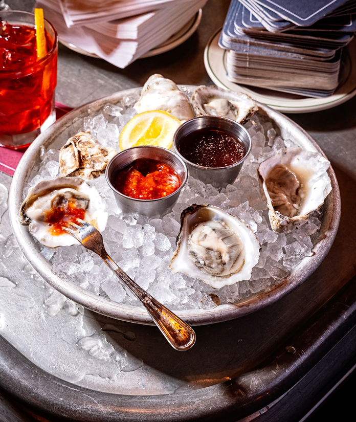 Fresh oysters on ice with lemon, sauces, and a drink, presented on a metal tray.