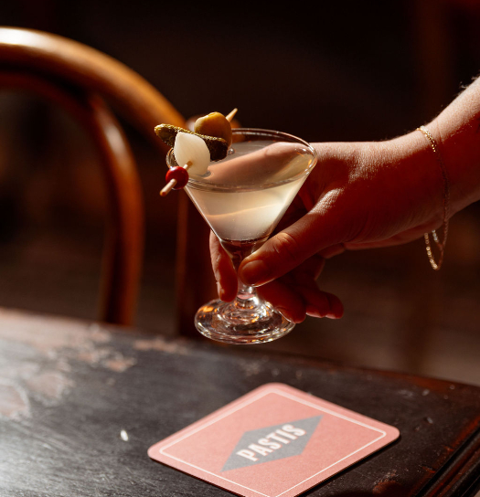 Hand holding martini glass with white cocktail and olive garnish above a table with menu card