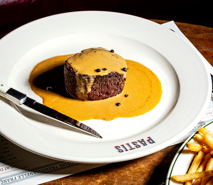 A filet mignon with peppercorn sauce on a Pastis restaurant plate, served alongside a side of fries.