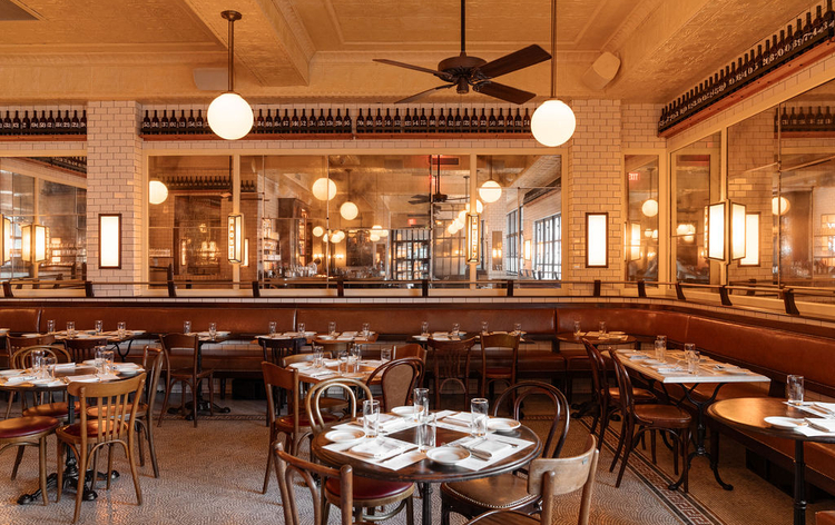 Cozy restaurant interior with wooden tables, chairs, and warm lighting. Large windows with framed artwork, ceiling fans, and tiled walls. ADA/WCAG compliant.