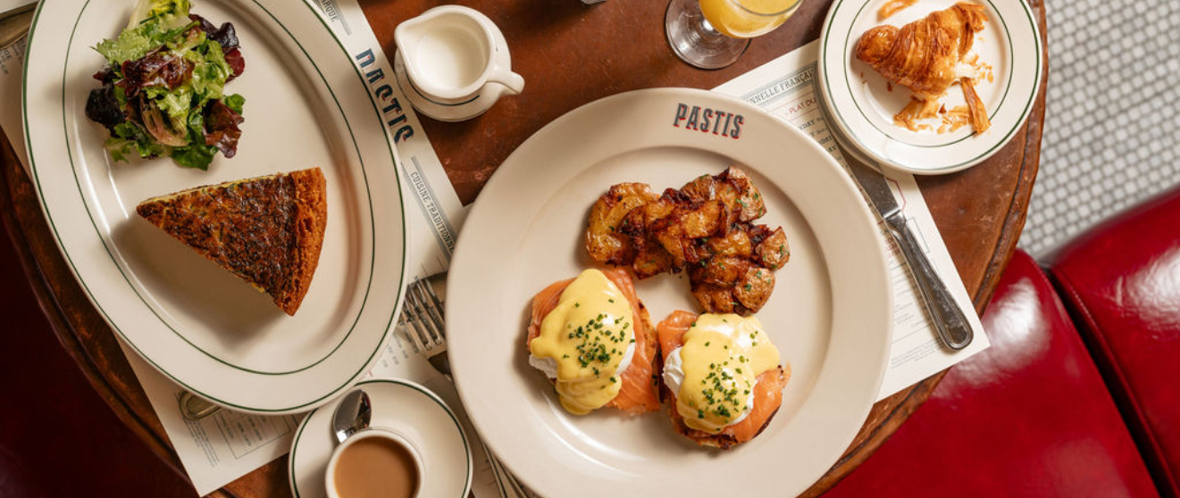 Overhead view of brunch dishes including eggs benedict with salmon, crispy potatoes, quiche with salad, and a croissant